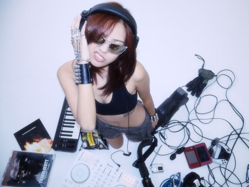 A woman in sunglasses and headphones surrounded by a DJ controller, keyboard, and tangled cables