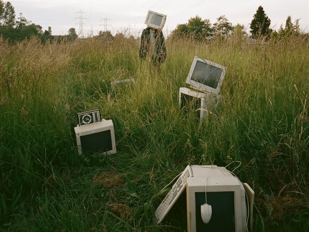 Old CRT monitors abandoned in a overgrown field, one worn like a mask by a figure standing among the grass