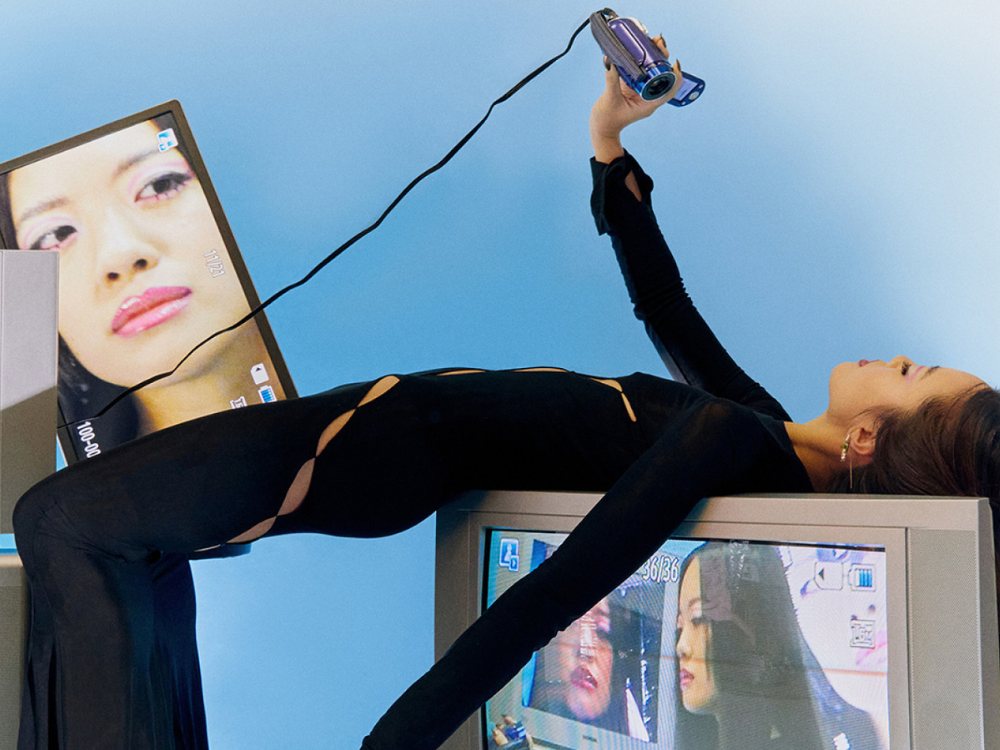A woman in black draped over a stack of vintage CRT televisions, filming herself with a small handheld camcorder