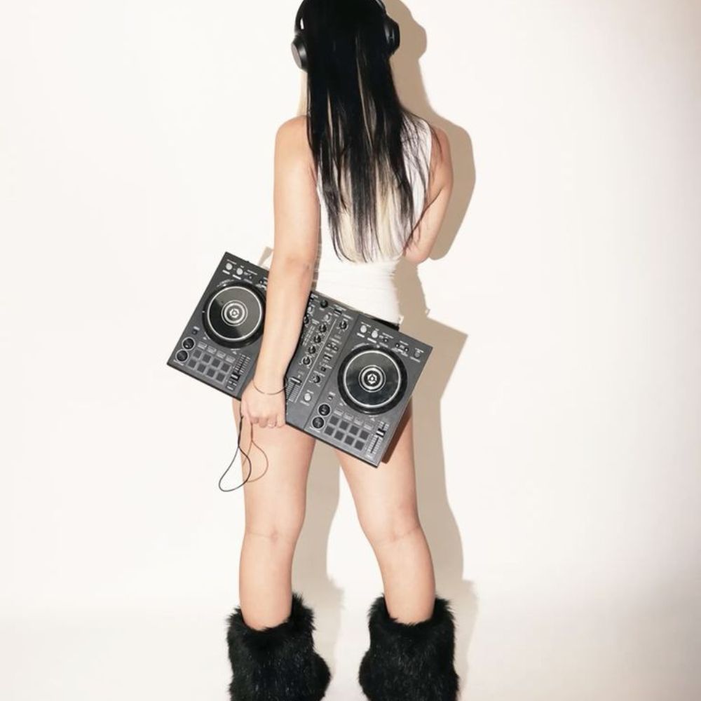 A woman seen from behind in a white tank top and furry black boots, holding two DJ controllers against her hips