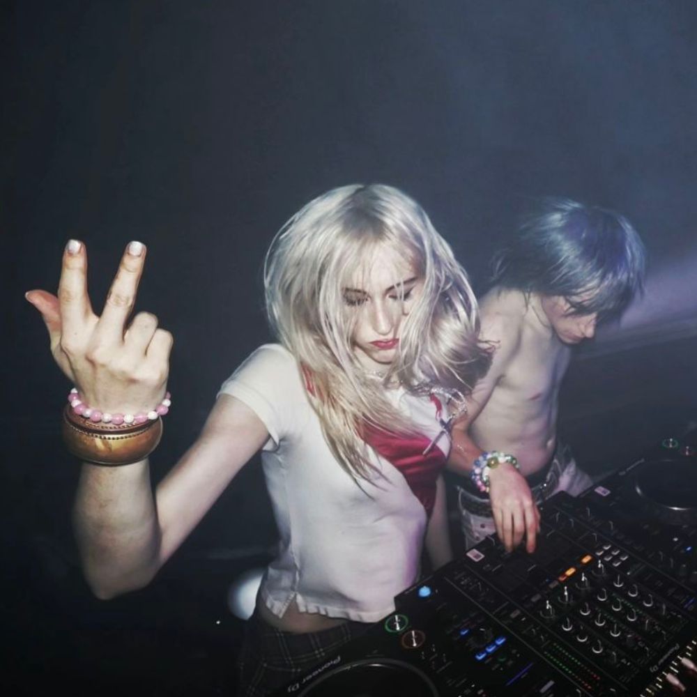 Two DJs at a dark, moody club booth, one throwing up a peace sign toward the camera